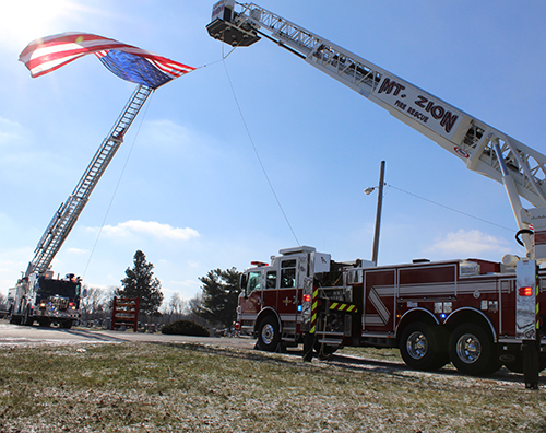 Photo by Mike B rothers Bethany Fire District Leads Procession  Sullivan, Lovington, Mt. Zion and area first responders joined family and friends to honor the passing of Jerry L. Clark, Chief Engineer of the Bethany Fire District. Following the 11 a.m. January 12 services at McMullin-Young Funeral Home in Sullivan the Bethany Fire District led Clark’s procession to Marrowbone Cemetery in Bethany where Sullivan and Mt Zion Fire District ladder trucks displayed the American Flag at the entry. Bethany American Legion Post #507 performed military rites. Serving the fire district for more than two decades, Clark was also a member of the Okaw Fire Association and Bethany American Legion Post.